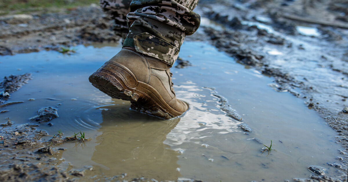 A close up of a person wearing military pants and brown boots trekking through a muddy puddle.